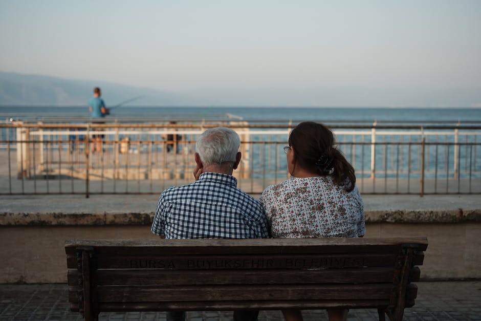 A elderly couple sitting on a bench on a waterfront, holding hands, looking happy and content with their lives in retirement.